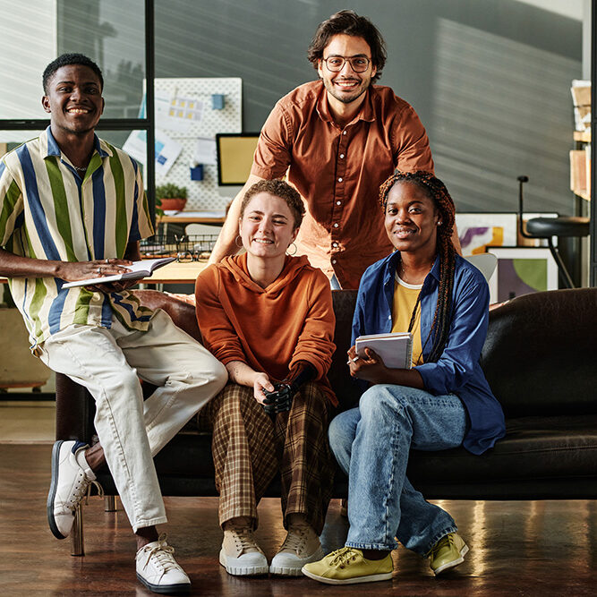 Four young successful interracial employees or creative designers in casualwear sitting on black leather couch in openspace office